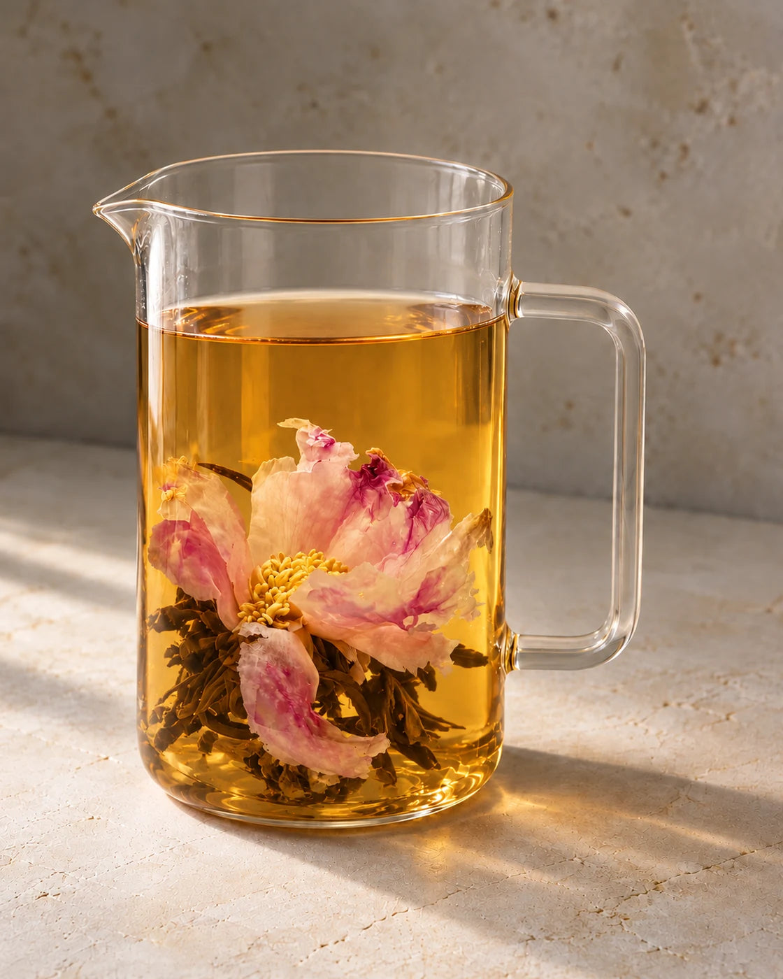 A pink peony unfurled above black tea leaves in amber tea inside a tall glass pitcher.