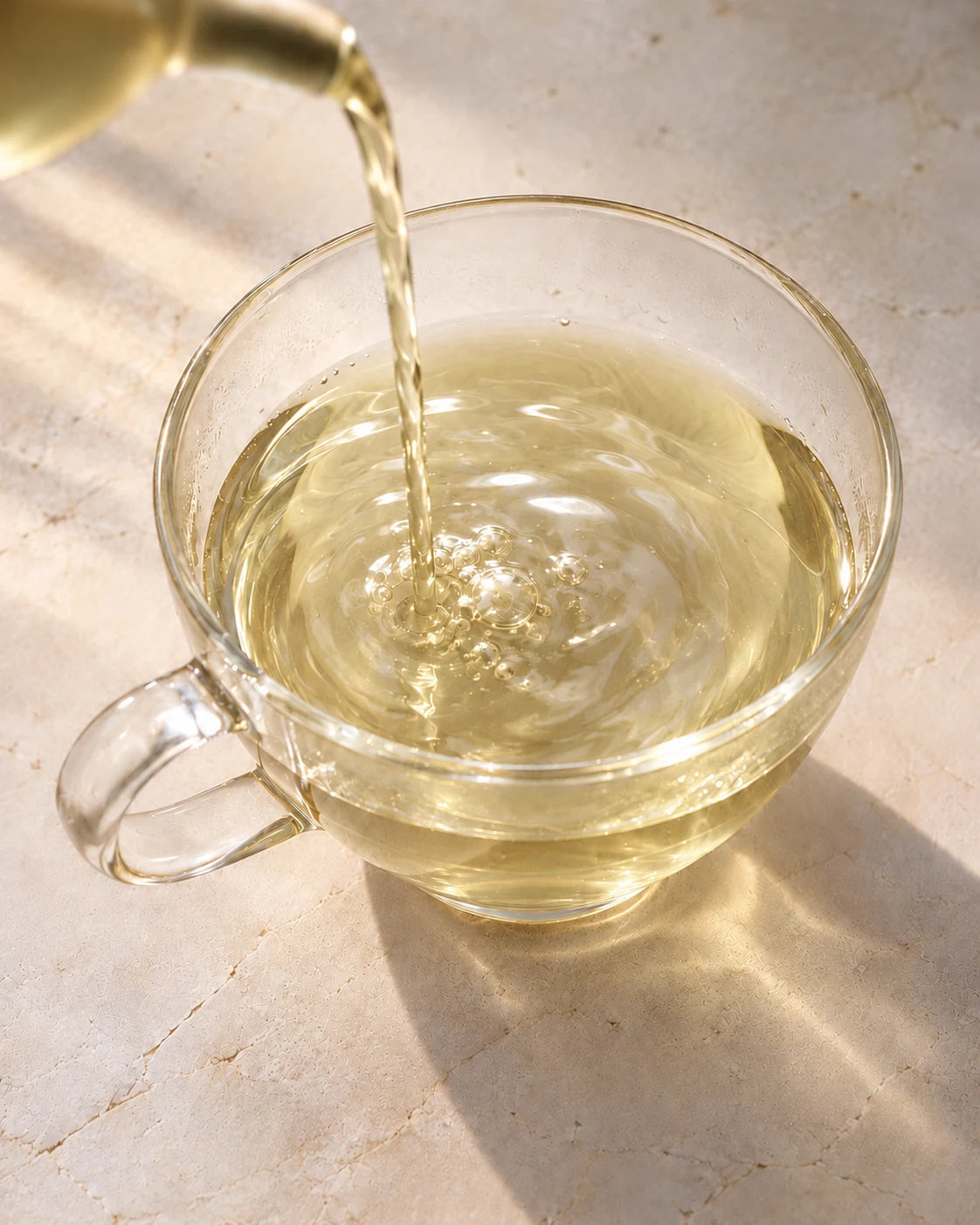 Clear, golden orchid green tea pouring into a glass cup, forming small bubbles and ripples.