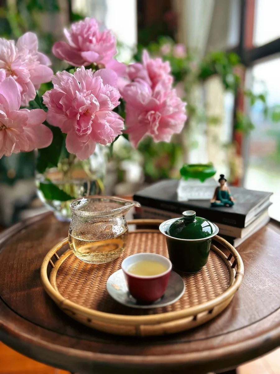 Tea served on a woven bamboo tray beside pink peonies and a sunlit window.