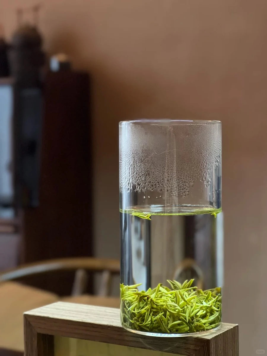 Mengding green tea leaves steeping in a tall glass on a wooden stand, with delicate condensation above the water.
