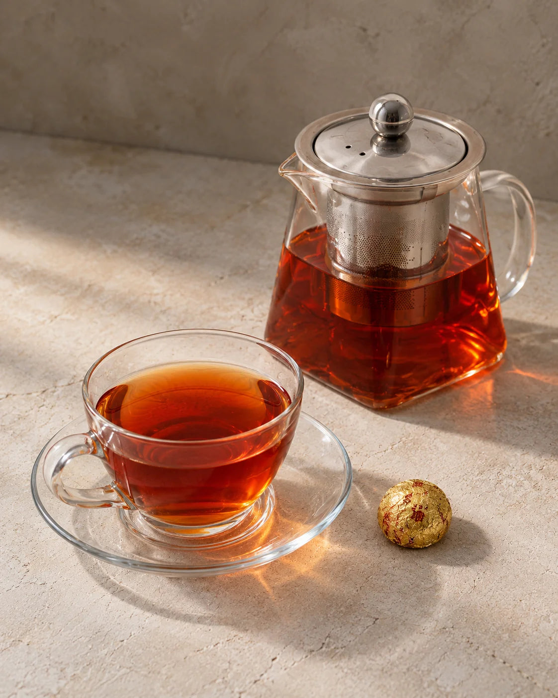 Deep ruby-red pu-erh tea in a glass teapot and matching cup, beside a gold-wrapped sticky rice mini tuo cha.