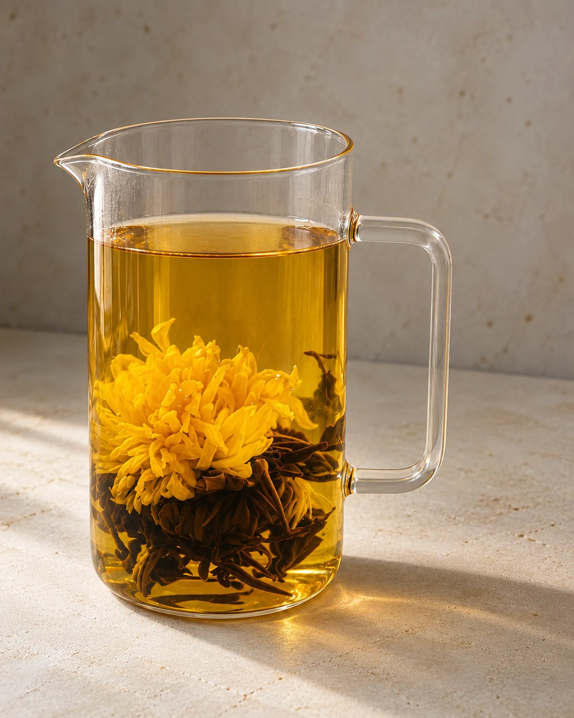 A golden chrysanthemum unfurled among black tea leaves in amber tea inside a tall glass pitcher.