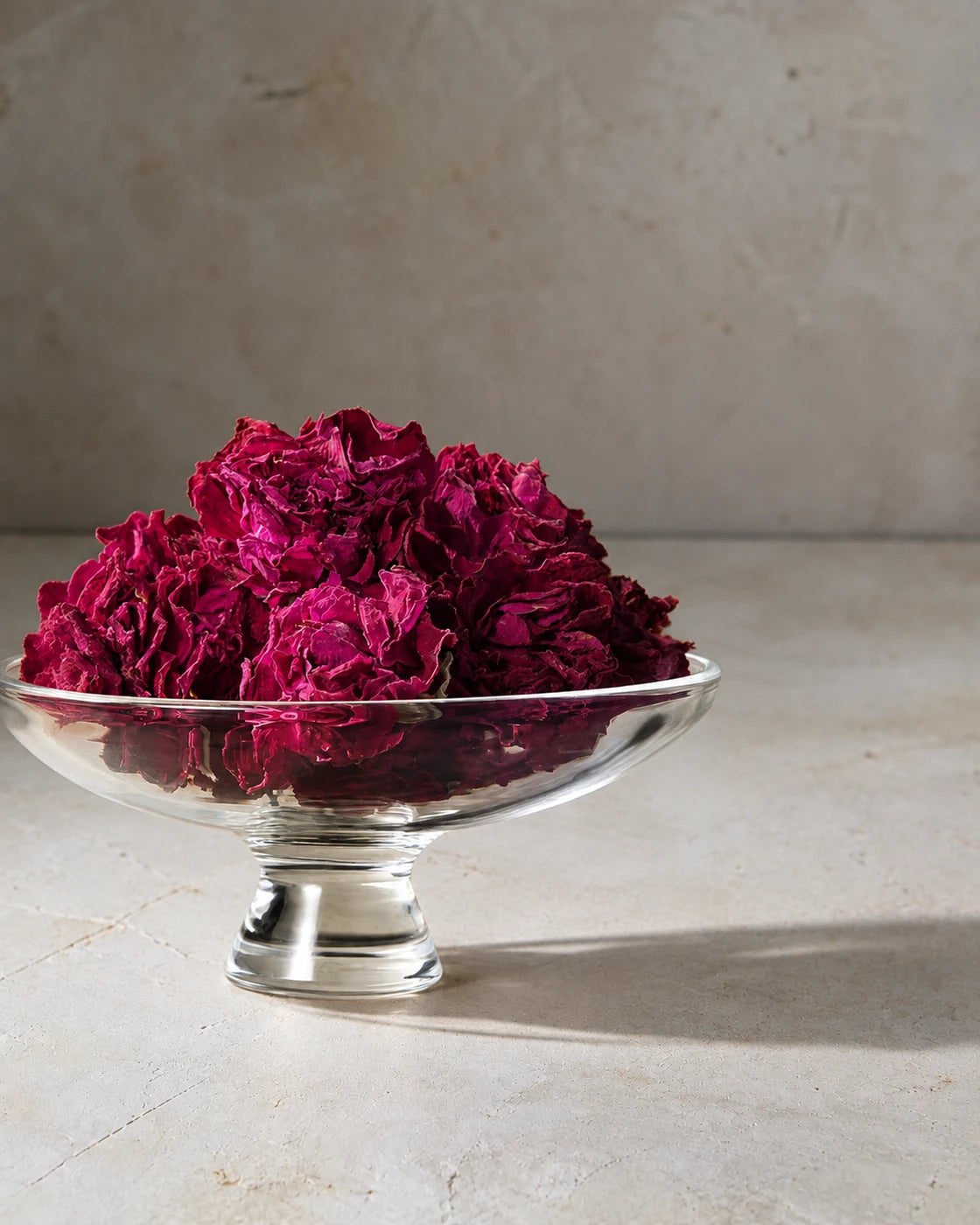 Whole dried rose flowers with deep pink petals piled in a clear glass pedestal dish.