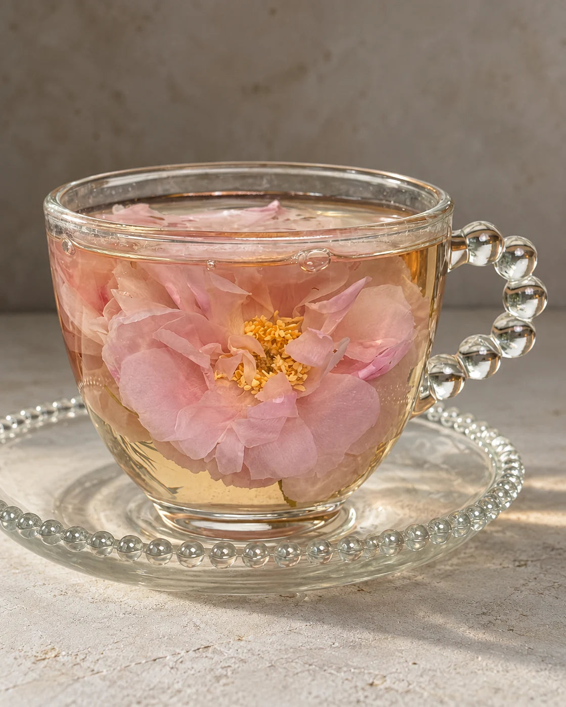 Pink rose blossoms steeping in pale golden tea in a glass cup with a beaded handle and matching saucer.