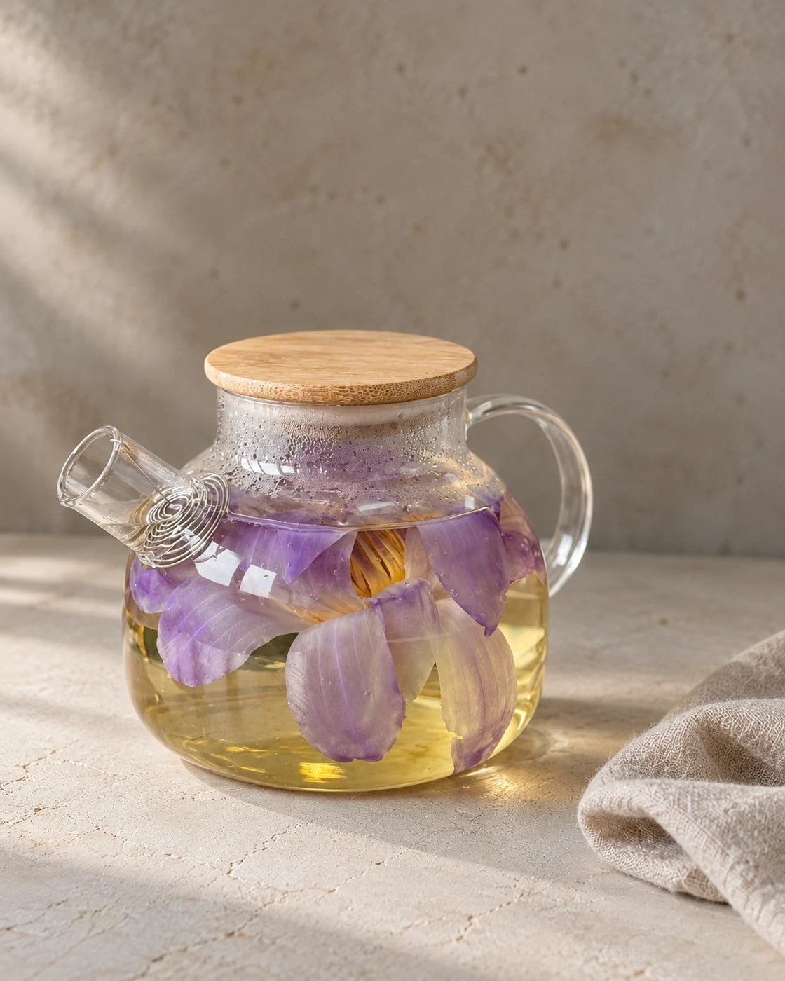 Blue lotus flower tea in a clear glass teapot with a wooden lid, showing purple petals in a golden infusion.