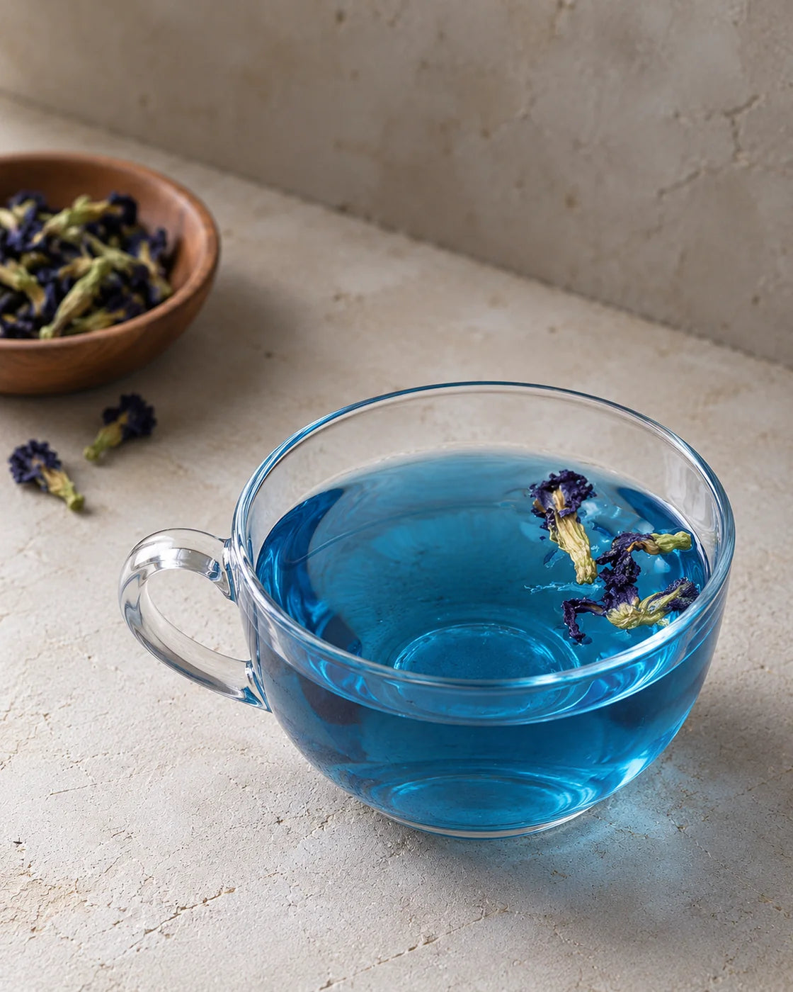 Blue butterfly pea flower tea in a clear glass cup with floating blossoms and a bowl of dried flowers behind it.