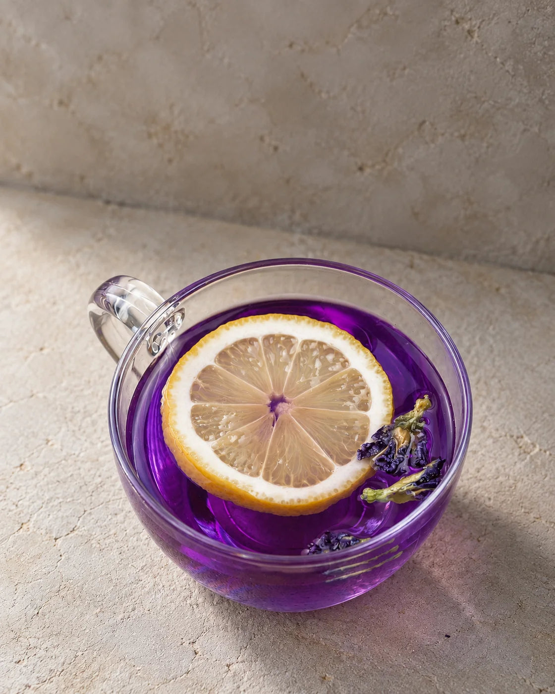 Purple butterfly pea flower tea in a clear glass cup with a lemon slice and floating blossoms.