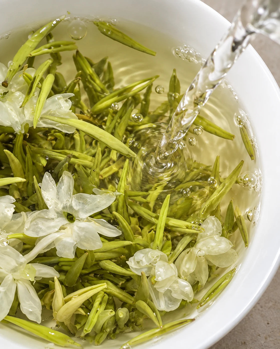 Water pouring over tender green tea buds and jasmine blossoms as they steep in a white bowl.