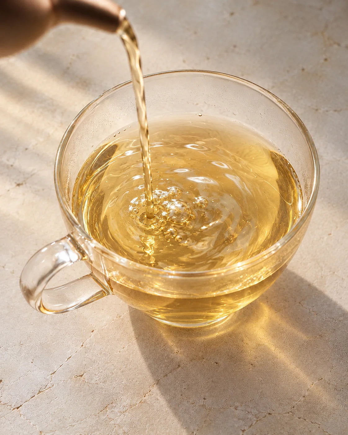 Clear, golden jasmine green tea pouring into a glass cup, forming small bubbles and ripples.