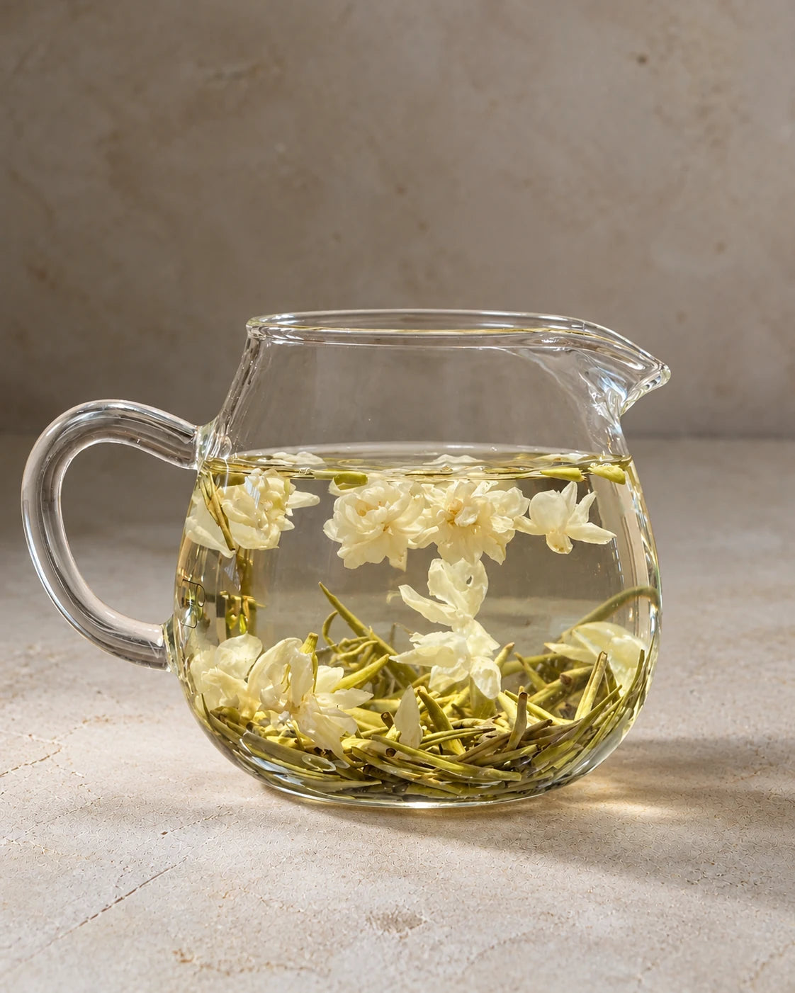 Jasmine green tea steeping in a clear glass pitcher, with floating jasmine blossoms and tea buds gathered below.