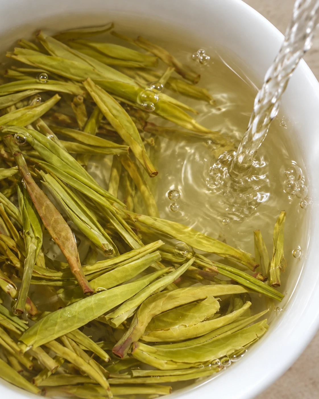 Water pouring over tender green tea buds as they steep in a white bowl, forming bubbles in the golden infusion.