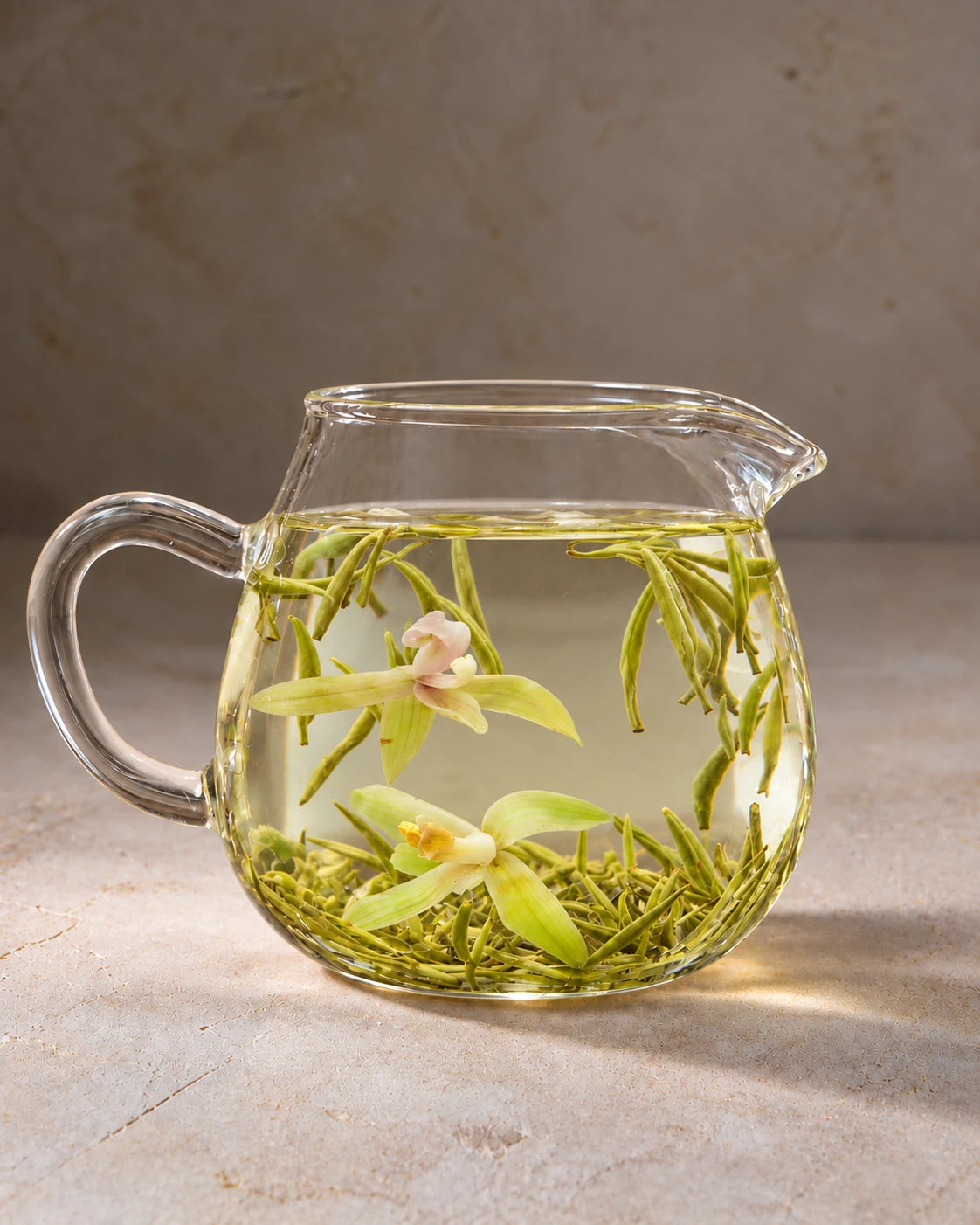 Orchid green tea steeping in a clear glass pitcher, with two orchid blossoms among slender tea buds in pale golden tea.