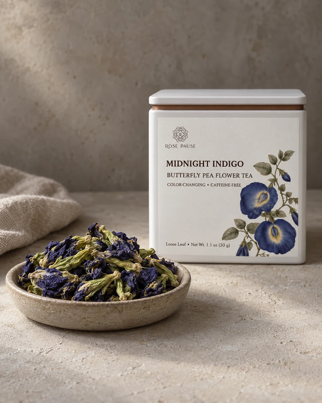 Rose Pause Midnight Indigo butterfly pea flower tea tin beside a ceramic dish of whole dried flowers.