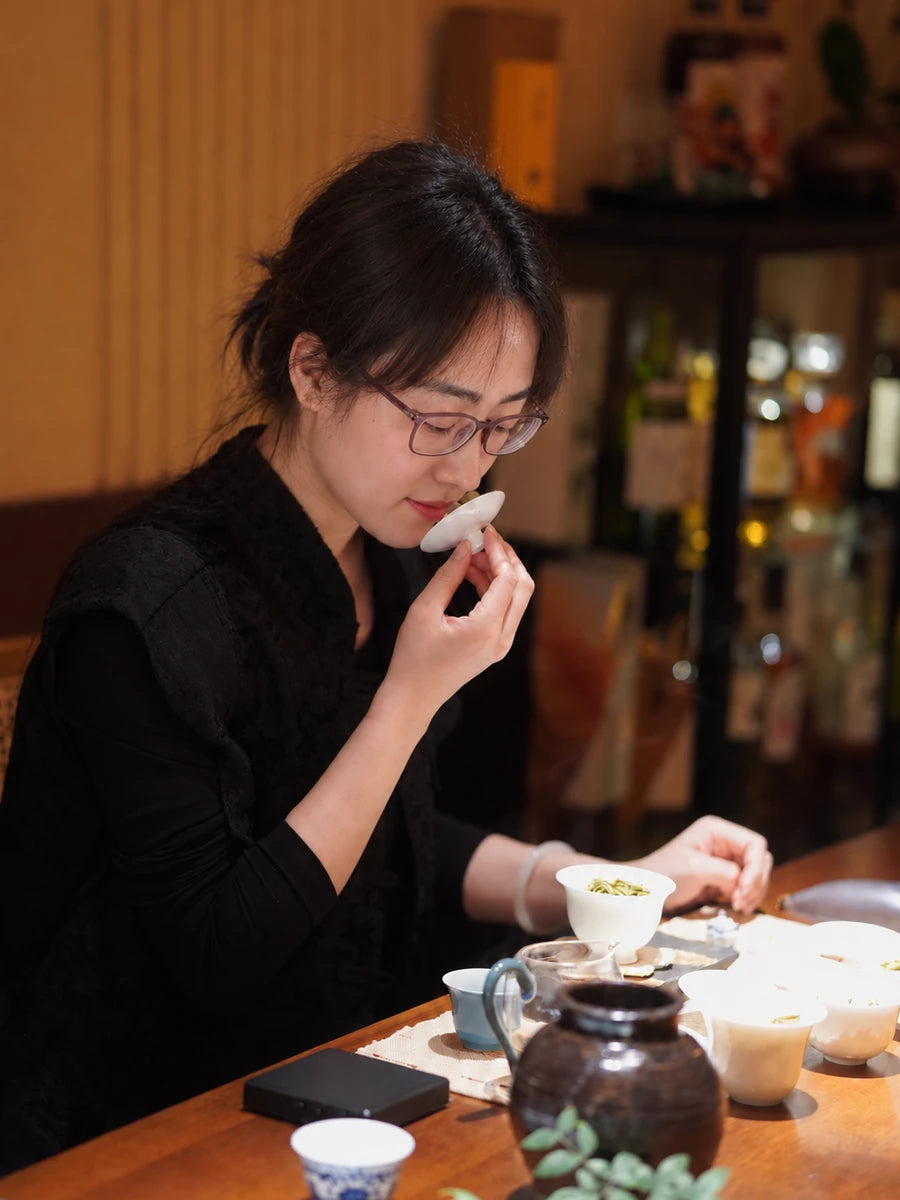 A woman appreciating the aroma from a white gaiwan lid at a wooden table set with tea and teaware.