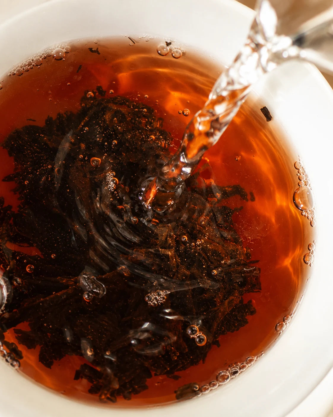 Water pouring over dark ripe pu-erh leaves in a white bowl, surrounded by reddish-amber tea and small bubbles.